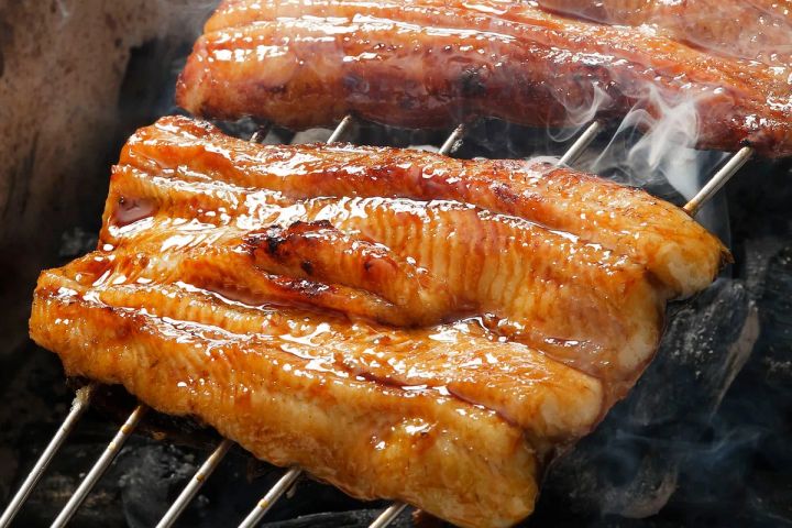 タレを塗りながら炭火で焼き上げられた鰻の蒲焼アップ写真。複数の鰻フィレットが金串に刺さり、煙が立ち上る焼き台の上でたれが照り光る調理工程のクロップショット