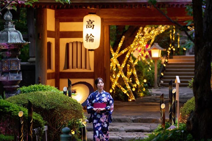 紺地に桜柄の浴衣姿の女性が「高輪」と書かれた提灯と竹あかりのイルミネーションが輝く日本庭園入口の石畳通路に立つ夜景写真