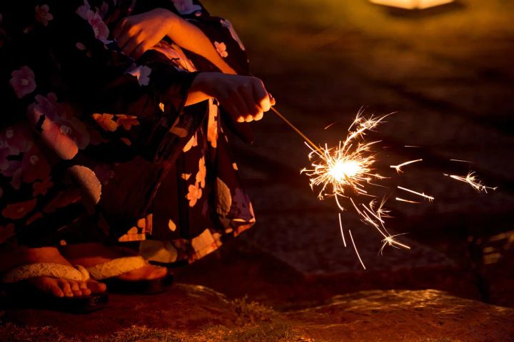 花柄の濃紺浴衣と下駄を着用した人物が夜の屋外で線香花火を手持ちしており、火花が広がる様子を暗闇の中で接写したショット