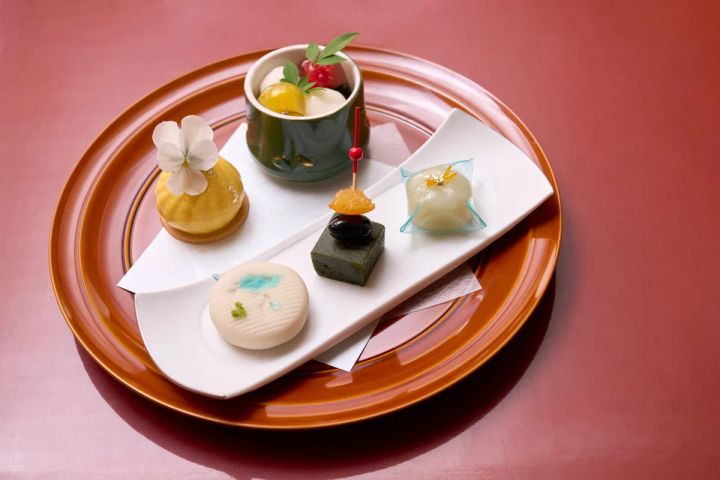 朱塗りの丸盆に和スイーツ5種を盛り付けた皿。抹茶羊羹・葛製和菓子・黄色いムース・白い落雁・竹筒入りフルーツゼリーが並ぶ夏の甘味プレート