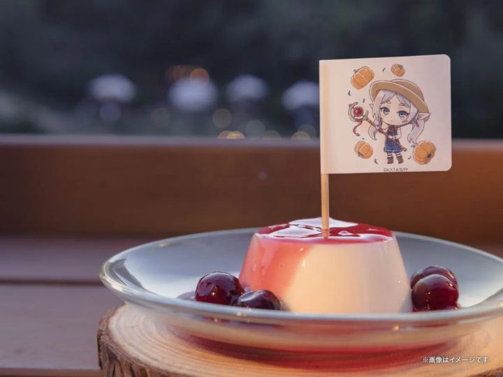 メルクーアプリン1個を屋外テラスで撮影した単体カット。フリーレンのちびキャラ旗が正面向きで大きく見え、背景は夕暮れの木立ちボケ。ガラス皿と木製コースターの組み合わせが確認できる