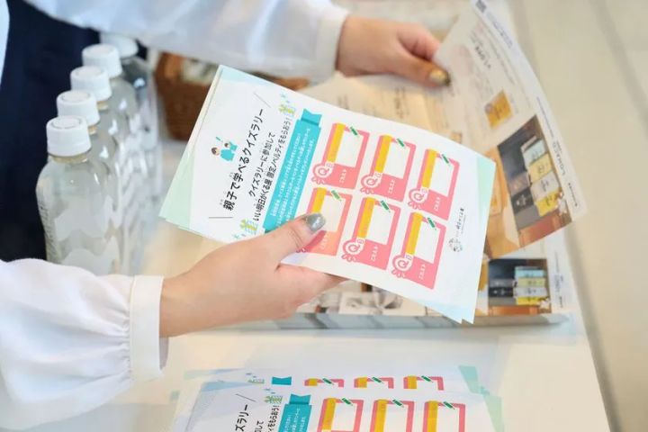 スタッフがクイズラリー参加シートを手に持つ様子。シートにはスタンプ欄6マスとアスクルキャラクターが印刷されており、会場受付台には水ボトルと展示パンフレットが並ぶ
