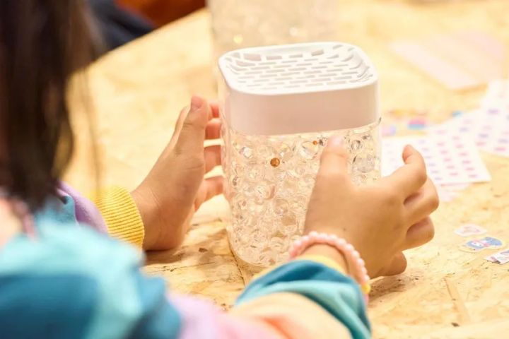 子どもがクリア容器型ゲル消臭剤をテーブル上で両手に持つ体験コーナーの様子。容器内に透明ビーズが入り、白いメッシュ蓋付きのプラスチック製容器を観察している