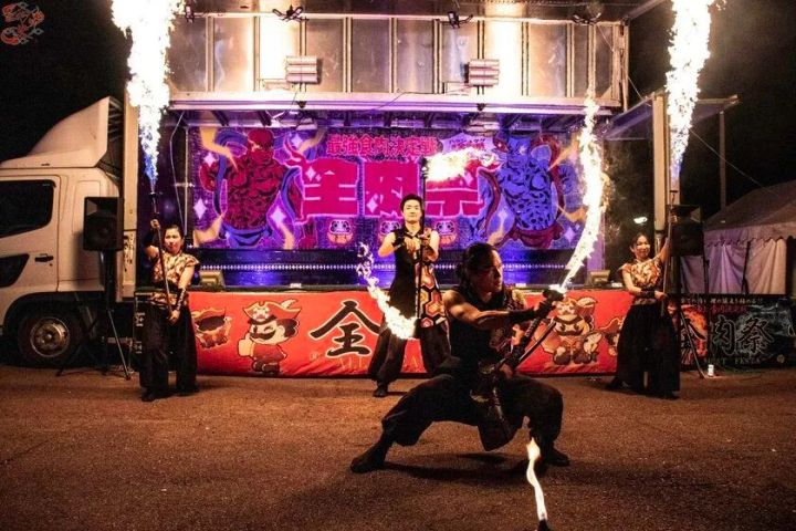 全肉祭ステージ前で黒装束の出演者4名がファイヤーパフォーマンスを披露している夜間ショットで、トラック荷台を改造したステージに全肉祭のロゴが照明で映し出されている