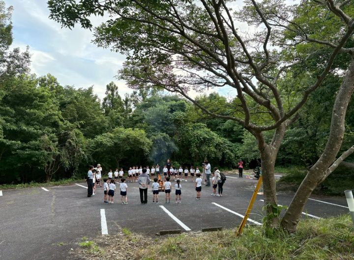 緑豊かな屋外駐車場で園児と思われる子どもたち約30名が整列し、引率スタッフとともに集団活動を行っている場面