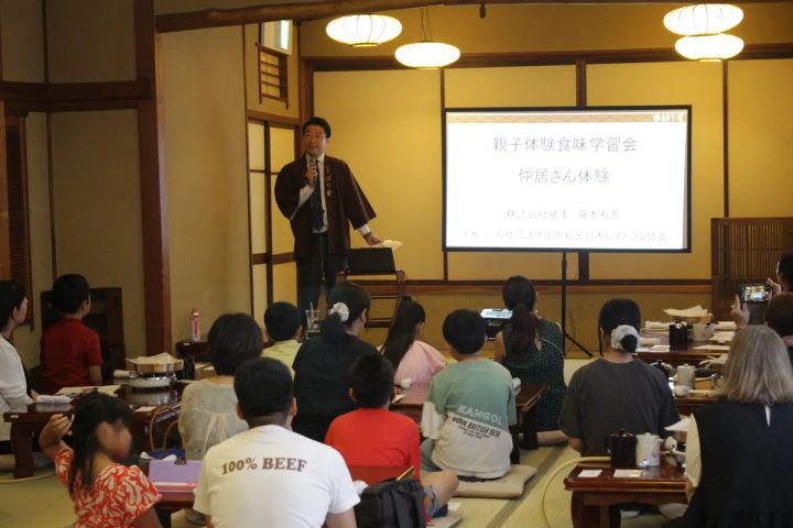 播重の和室座敷で株式会社播重代表が親子体験食味学習会・仲居さん体験のスライドを映しながら小中学生と保護者約20名に向けて講義している様子