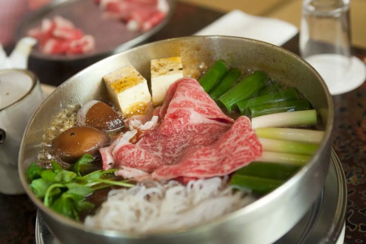 すき焼き鍋のクローズアップ。霜降り牛肉スライス・豆腐・椎茸・長ねぎ・春菊・糸こんにゃくが醤油ベースのだし汁で煮られている調理中の状態。奥に生肉の入った器も写る。
