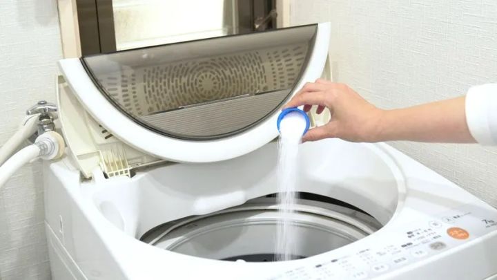 縦型全自動洗濯機の投入口に、付属の計量カップで白色粉末洗剤を注ぎ入れている使用シーン写真。オキシクリーン粉末タイプの実際の使い方を示す