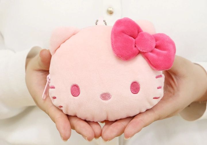 濃いピンクのリボンが付いたピンクのハローキティ顔型ぬいぐるみポーチを両手で持つ、白い服の女性