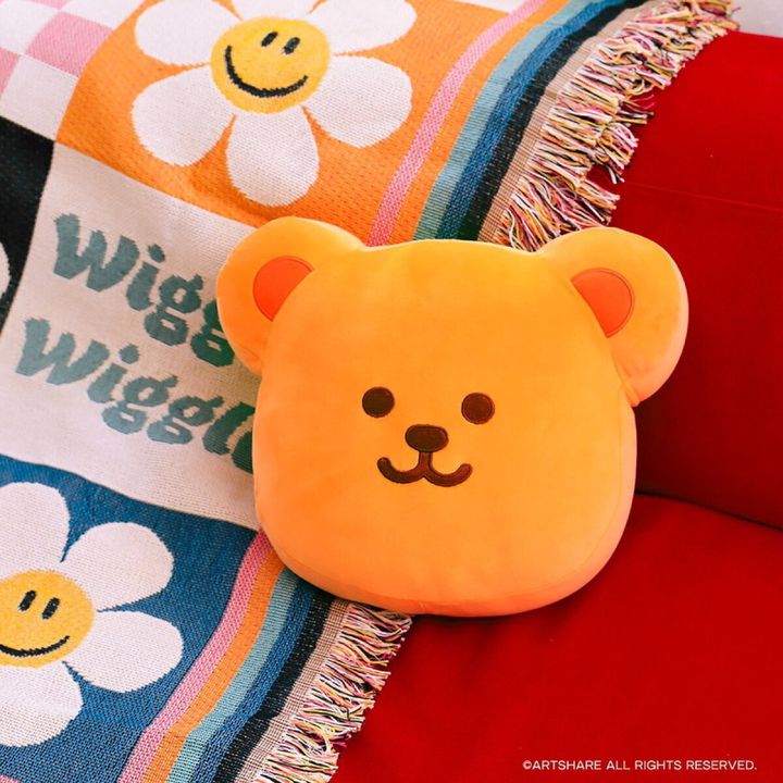 wiggle wiggleのウィグルベアをかたどったオレンジ色のもちもちクッション。赤いソファの上に置かれ、背景にスマイル花柄でWiggle Wiggleとプリントされたフリンジ付きブランケットが見える