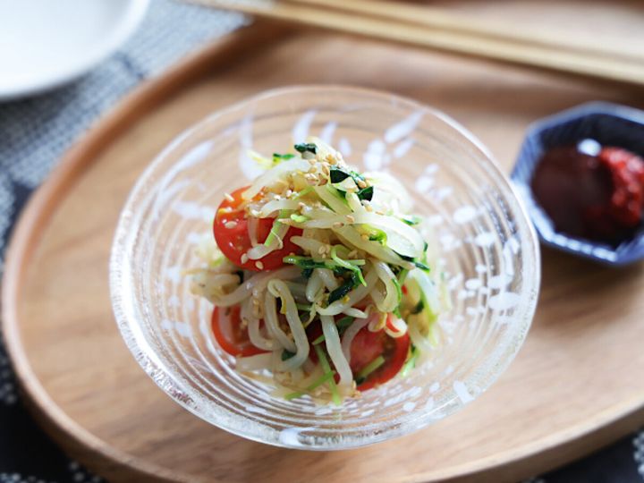 ガラスの器に盛られたもやしとプチトマトの無限ナムル。半分に切ったプチトマト・豆苗・白ごまがのり、木製トレーの奥に赤い薬味を入れた小皿と箸が置かれている
