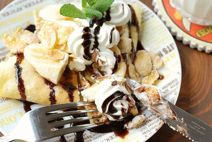 文字柄の皿に盛られた、ホイップクリームとスライスバナナ、チョコソース、コーンフレークをのせたクレープをフォークとナイフで切り分ける手元アップ