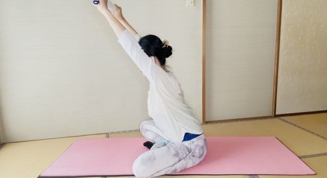 座った姿勢でタオルを両手に持ち、腕を斜め上に伸ばして背中を伸ばす女性