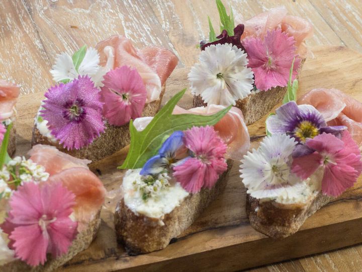 バゲットに生ハムとピンク・紫・白のエディブルフラワー、緑の水菜を彩りよく乗せた完成したタルティーヌを斜めから撮影した写真