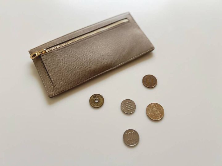 白いテーブルの上に置かれたブロンズ色の薄型長財布と、5円・10円・100円などの硬貨が数枚並んでいる