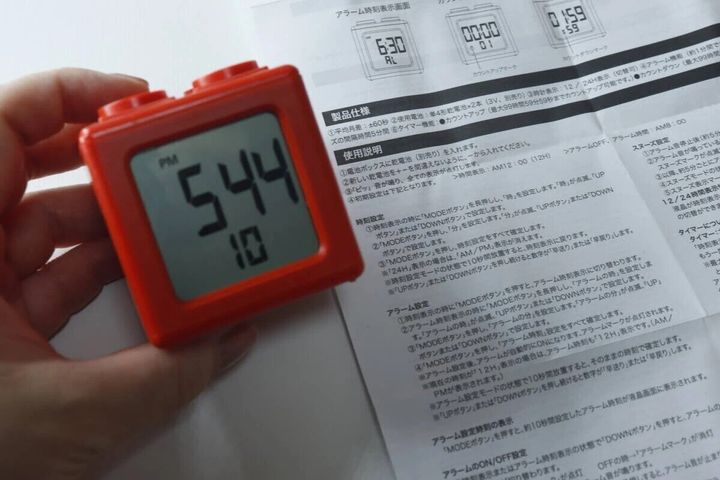 赤いブロック型デジタルタイマー時計を手に持ち、時刻表示「5:44」が見える。横には説明書