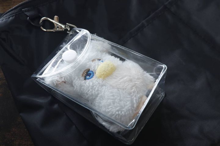 黒いバッグの上に、白い毛のぬいぐるみマスコットが入った透明なポーチが置かれている