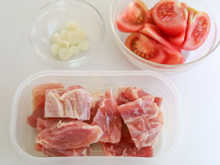 白いテーブルに並んだ、うす切りにしたにんにく・くし切りにしたトマト・一口大に切った生の鶏もも肉の下準備の様子