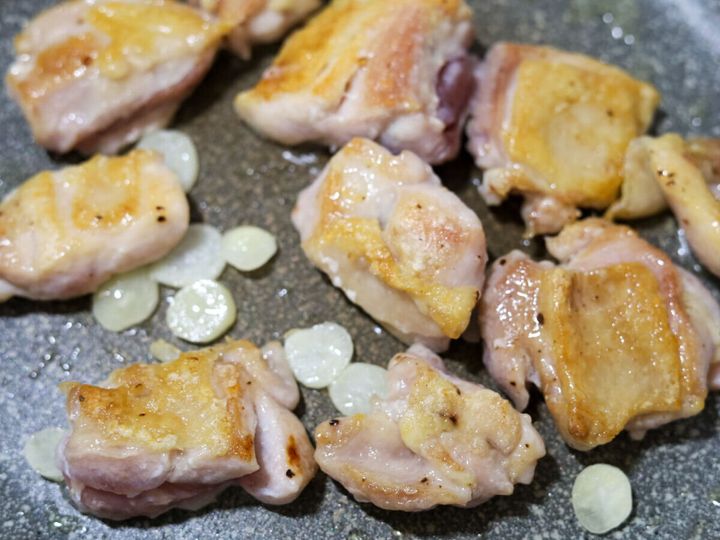 フライパンでこんがり焼き色がついた鶏もも肉を裏返し、うす切りにんにくを加えて焼いている様子