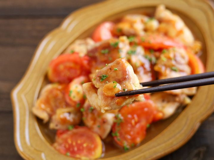 茶色い楕円形の器に盛られた鶏肉とトマトのガリバタ炒めを、黒い箸で鶏もも肉を一切れつまみ上げている様子