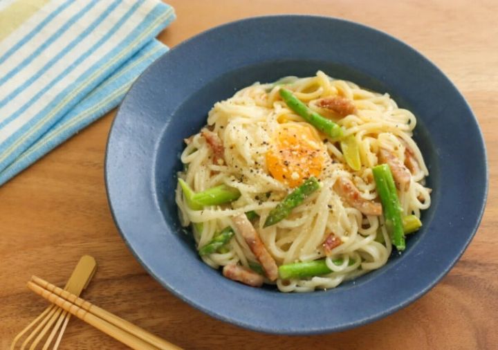 紺色の器に盛ったクリーミーなカルボナーラ風うどんに、アスパラガス・厚切りベーコン・卵黄をのせ黒こしょうをふった写真