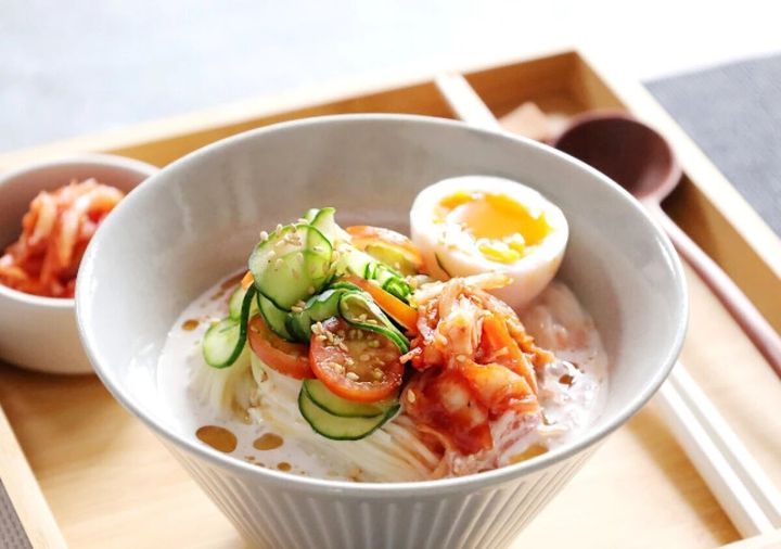 白い器に盛ったそうめんと豆乳スープのコングクスに、きゅうり・プチトマト・キムチ・半熟卵をのせ白ごまをふった写真