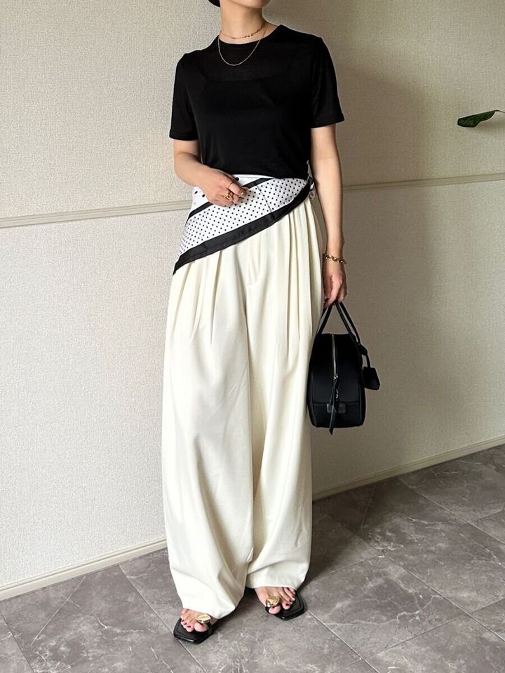 黒いシアーTシャツと白いワイドパンツを着用し腰に水玉スカーフを巻いた女性が立っている