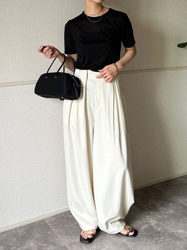 黒いシアーTシャツと白いワイドパンツを着用し黒いバッグを持った女性が立っている