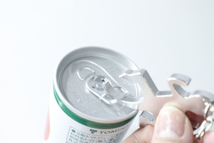 人の指が銀色のトカゲ型栓抜きキーホルダーで缶ジュースのプルタブを起こそうとしている
