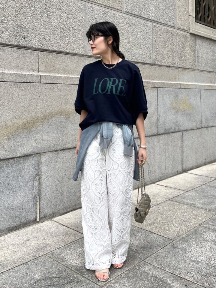 ネイビーのロゴTシャツと白いレースパンツを着用し、腰にシャツを巻いた女性