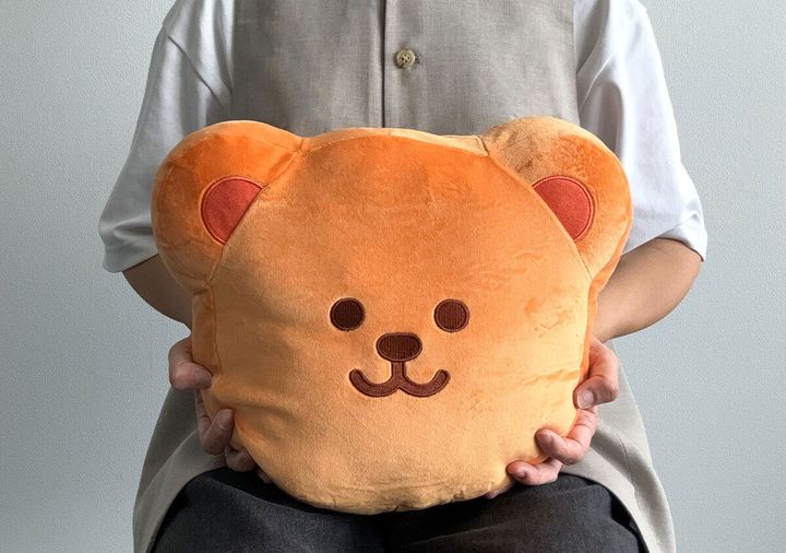 白いTシャツとベージュのベストを着た人が、ウィグルベアのもちもちクッションを抱きかかえている