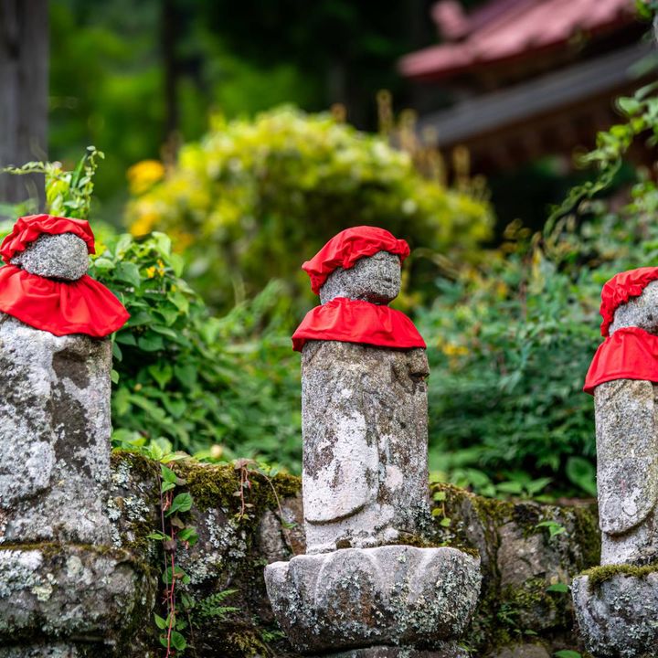 赤い前掛けと帽子をつけた、三体のお地蔵さんが並ぶ田舎道の風景