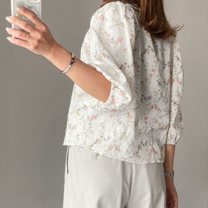 白い花柄ブラウスとベージュパンツを着た女性の後ろ姿。スマホで撮影中