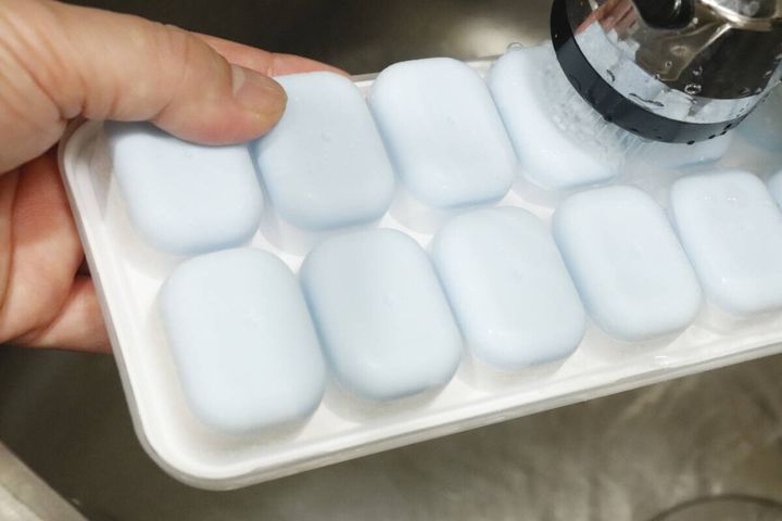 凍った氷が入ったアイストレーの底に、蛇口から出るシャワーで水をかけている手のアップ