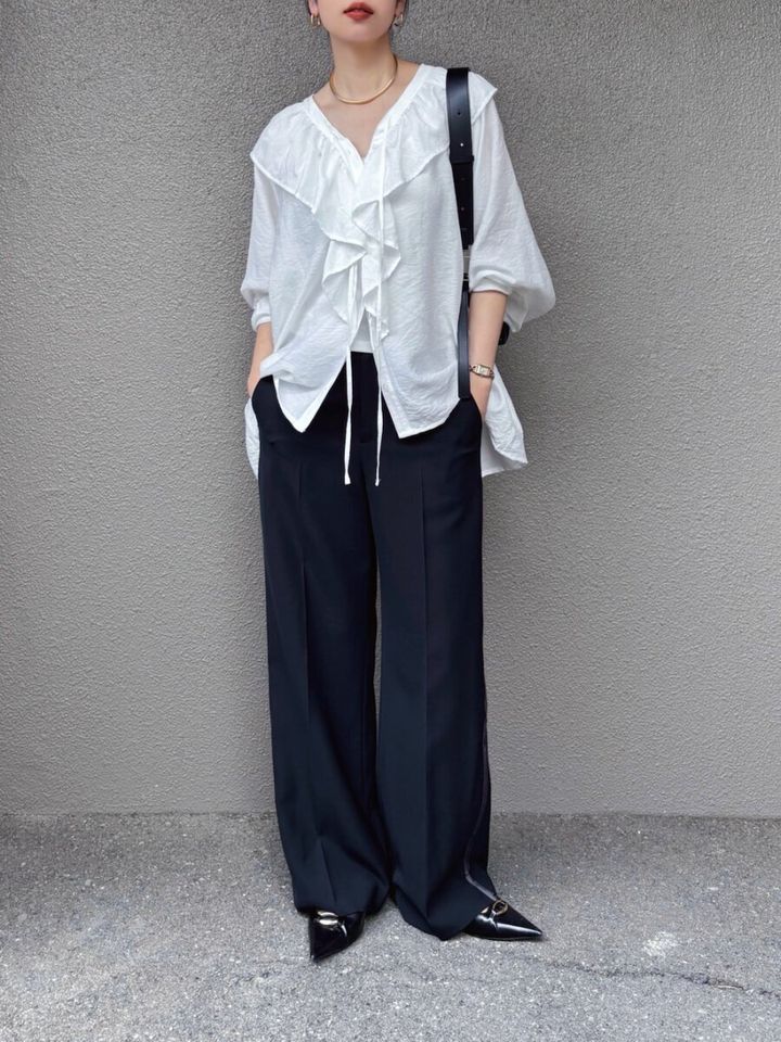 白いフリルブラウスと黒ワイドパンツの女性が立つ