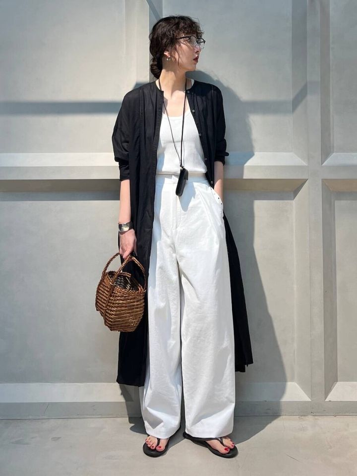 白いタンクトップとワイドパンツに黒のボタン付きワンピースを羽織り、カゴバッグとサンダルを合わせた女性の全身