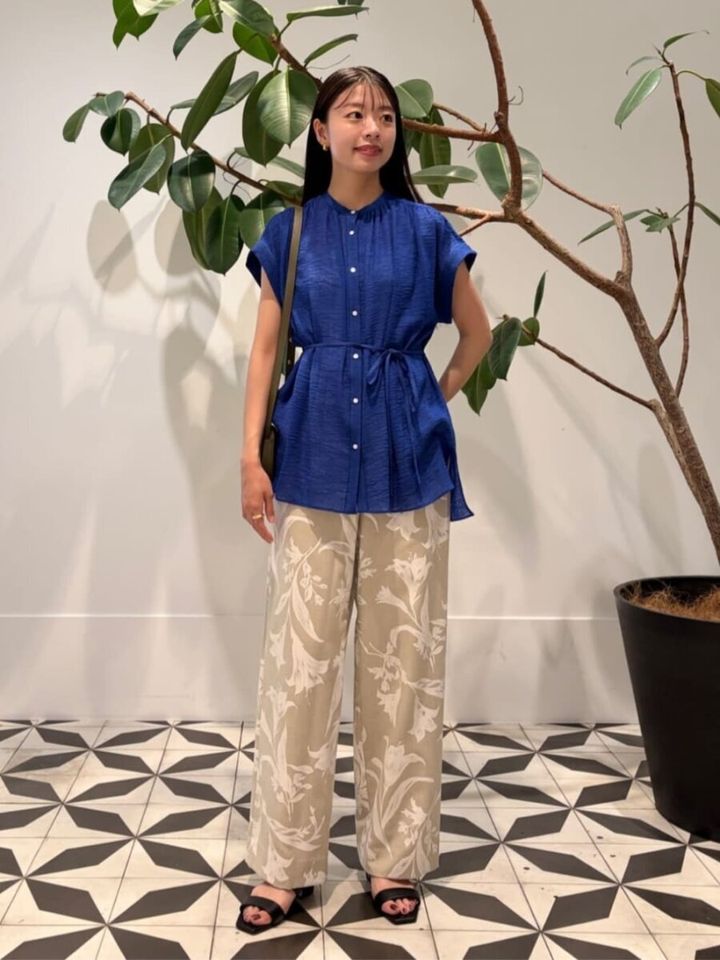 青いブラウスと花柄パンツの女性。黒サンダルを履き、観葉植物の横に立つ。