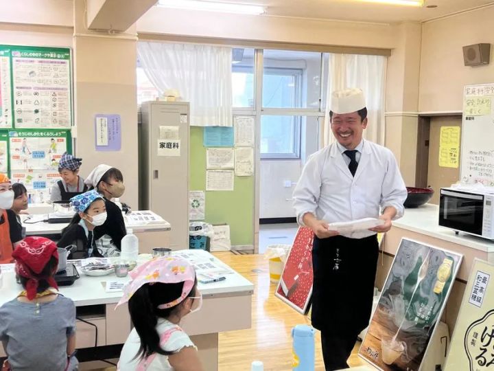 白衣と帽子、黒い前掛けを身につけた講師が、家庭科室で子どもた