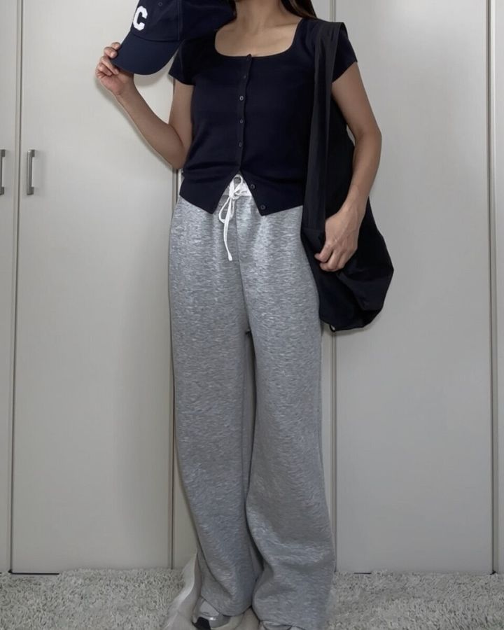 ネイビーのカーディガンとグレーのスウェットパンツ、キャップを持った女性