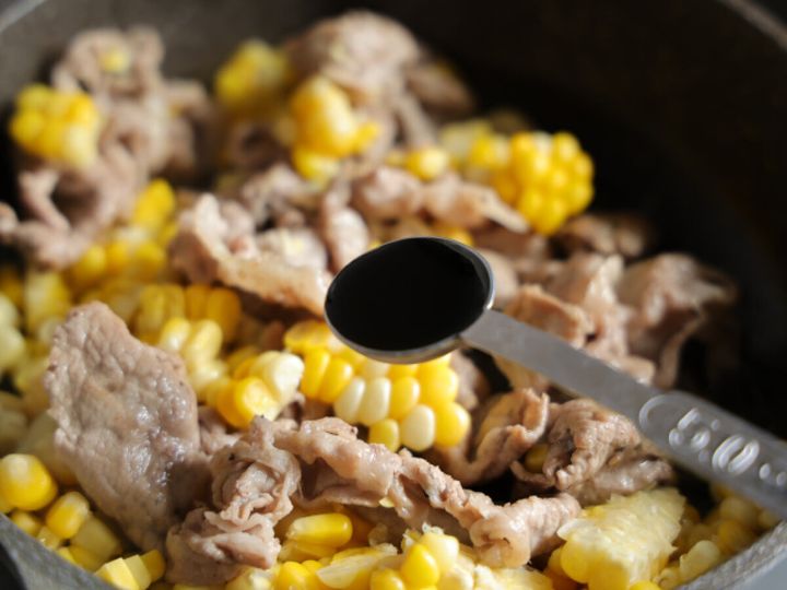 炒めている豚肉ととうもろこしが入ったフライパンに、計量スプーンで醤油を回し入れている。