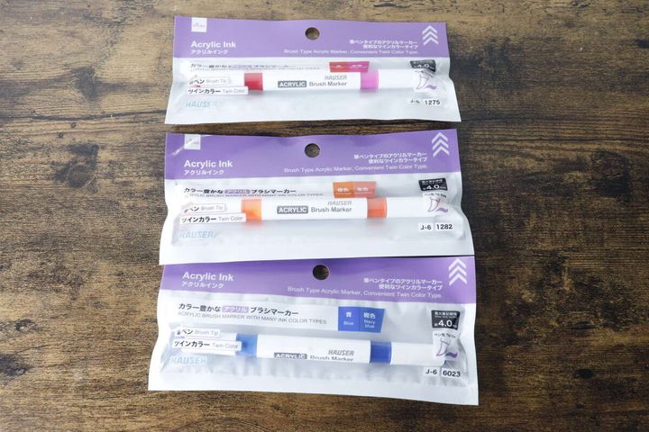 3つのアクリルブラシマーカーがパッケージに入った状態で木製のテーブルに置かれている