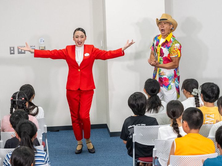 会場前方で赤いスーツの「綾小路麗華」とアロハ姿のMCが腕を広げて登場する様子