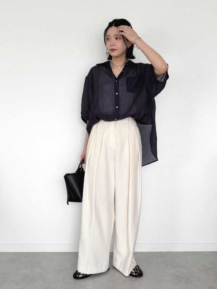 黒のシアーシャツと白のタックワイドパンツを着て髪に手を添える女性
