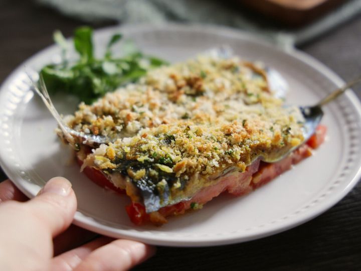 白いお皿に盛り付けられた、焼き上がったいわしとトマトのパン粉焼き。手前で皿を持っている手元