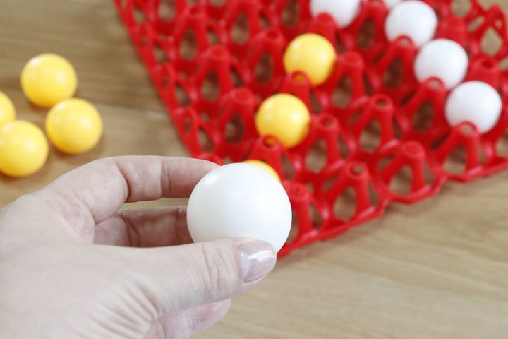 女性の手が白いピンポン玉を一つ持ち、ボードに置かれた玉を見ている