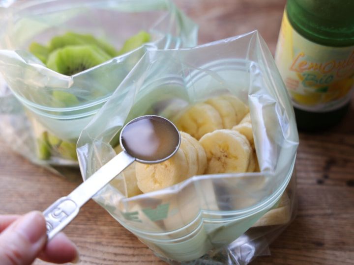スプーンで計量したはちみつを、輪切りバナナが入った密閉袋に加えている手元。奥にはレモン汁のボトルが見える。