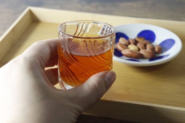 ミニグラスに入った琥珀色の飲み物を持ち、奥におつまみ