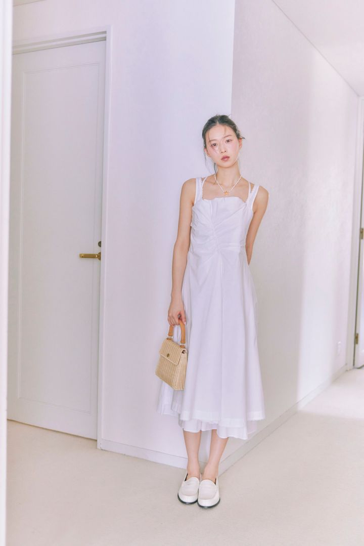 白い壁の室内で、白のキャミソール風ワンピースとローファーを身にまとい、カゴバッグを手にする片山友希