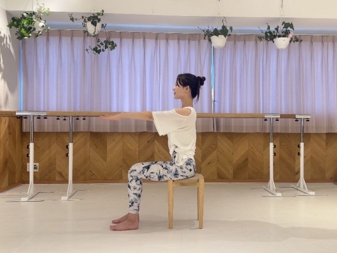 椅子に浅く座り、両手を前へ伸ばし、息を吸いながら背筋を伸ばす。