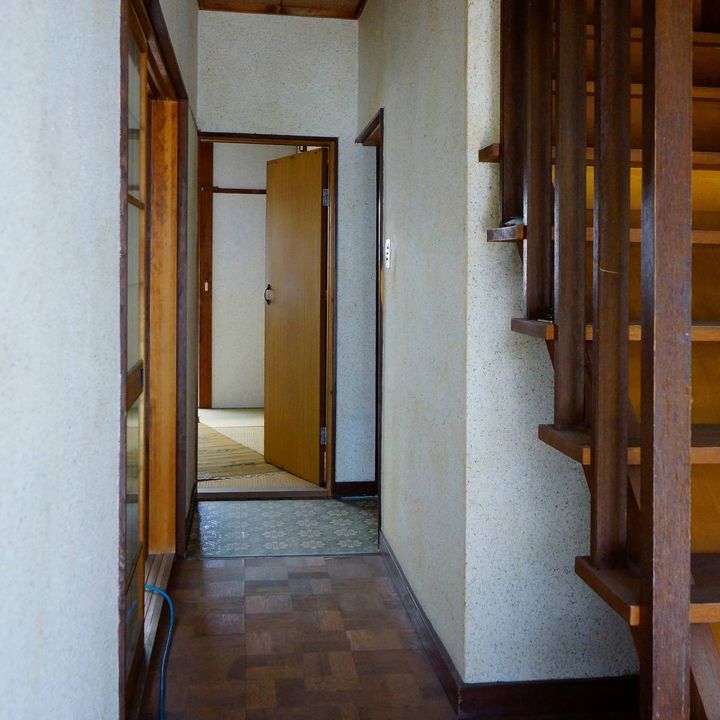 古い木造住宅の玄関。右手に階段があり、奥へ続く廊下と開いた室内ドアが見える。